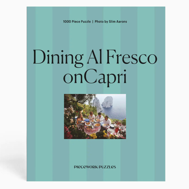 Part of the Slim Aarons Puzzle Collection by Piecework, this 1000-piece jigsaw puzzle titled “Dining Al Fresco on Capri” features a group dining outdoors by the sea with distant cliffs, set against a striped teal background. Crafted from high-quality art paper, it offers vibrant colors and lasting enjoyment.