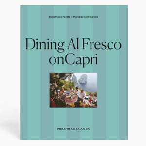 Part of the Slim Aarons Puzzle Collection by Piecework, this 1000-piece jigsaw puzzle titled “Dining Al Fresco on Capri” features a group dining outdoors by the sea with distant cliffs, set against a striped teal background. Crafted from high-quality art paper, it offers vibrant colors and lasting enjoyment.