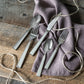 The Simon Pearce Woodstock 5-Piece Flatware Setting—two forks, two spoons, and a knife—is displayed on rustic wood with a purple napkin and twine, ideal for modern tables.