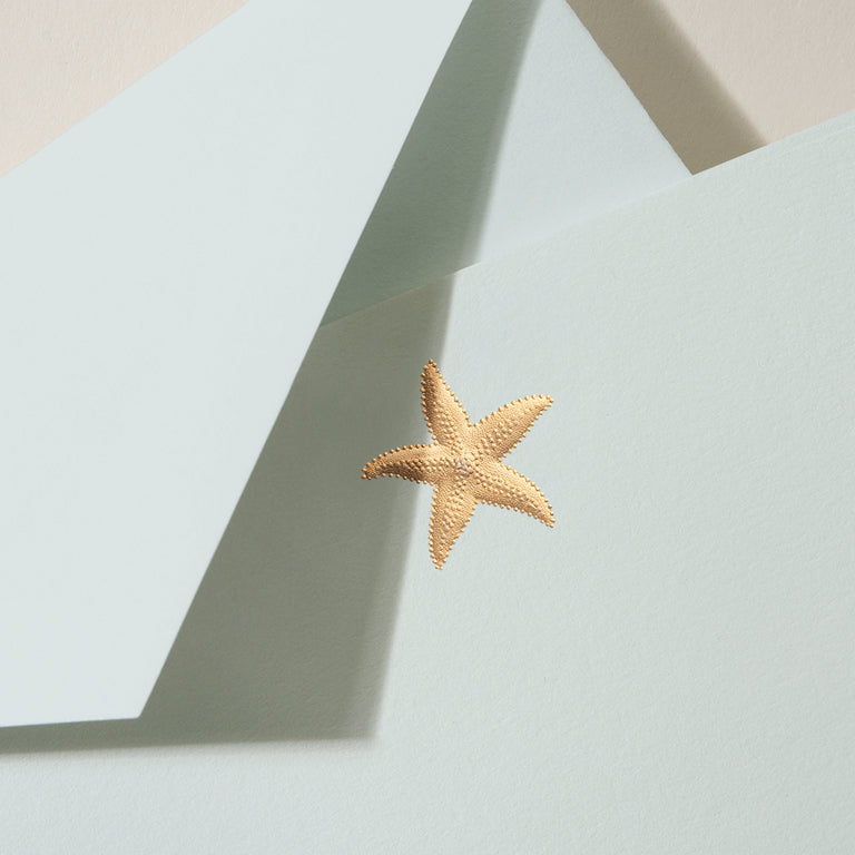 A gold starfish sticker decorates a light blue envelope layered over another blue one, echoing a nautical vibe. Ideal for beach-themed stationery or pairing with the Crane Gold Starfish Beach Glass Flat Card Set.