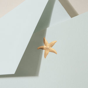 A gold starfish sticker decorates a light blue envelope layered over another blue one, echoing a nautical vibe. Ideal for beach-themed stationery or pairing with the Crane Gold Starfish Beach Glass Flat Card Set.