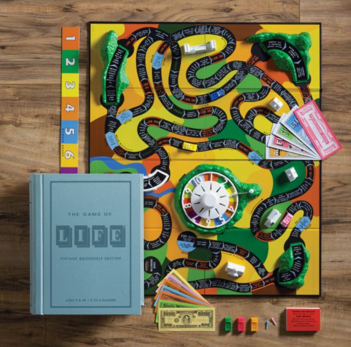 The image showcases The Game of Life Vintage Bookshelf Edition by WS Game Company set up with the board, cards, money, spinner, and game pieces neatly arranged for play. A closed game box and instruction booklet are placed beside the board, adorned with vintage graphics that add a touch of nostalgic charm.