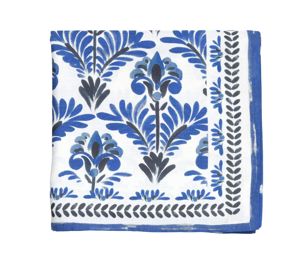 The Botanica Tablecloth Collection by Kim Seybert features a white cloth napkin with a blue and black floral pattern and a blue border, evoking a vintage tablecloth style.