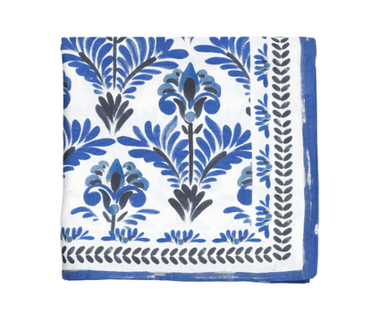 The Botanica Tablecloth Collection by Kim Seybert features a white cloth napkin with a blue and black floral pattern and a blue border, evoking a vintage tablecloth style.