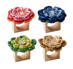 Four Kim Seybert Pareo Napkin Rings, each topped with a hand-crocheted flower in pink, blue, green, or tan, are arranged in a grid on a white background.