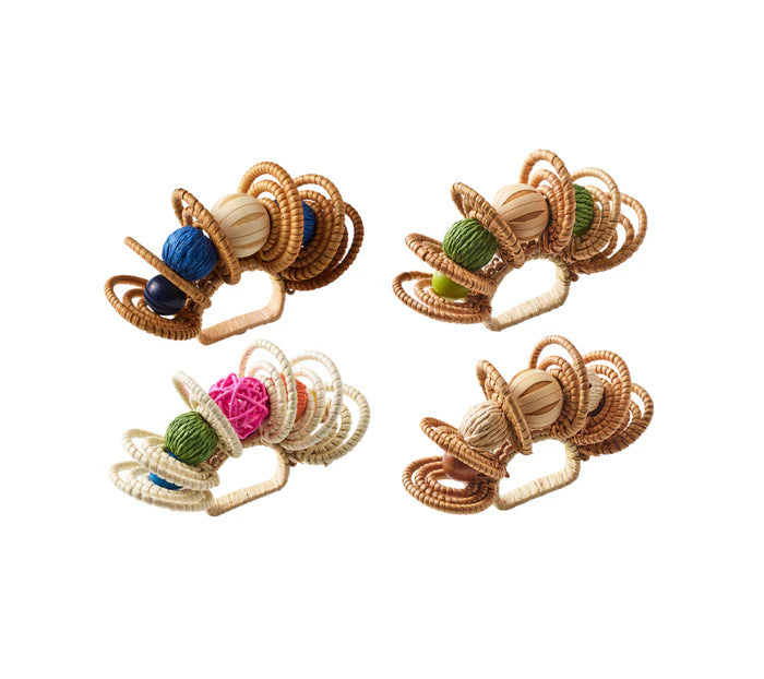 Four Calypso Napkin Rings by Kim Seybert, featuring woven rattan, hand-dyed wooden beads, and colorful yarn balls, artfully arranged on a white background.