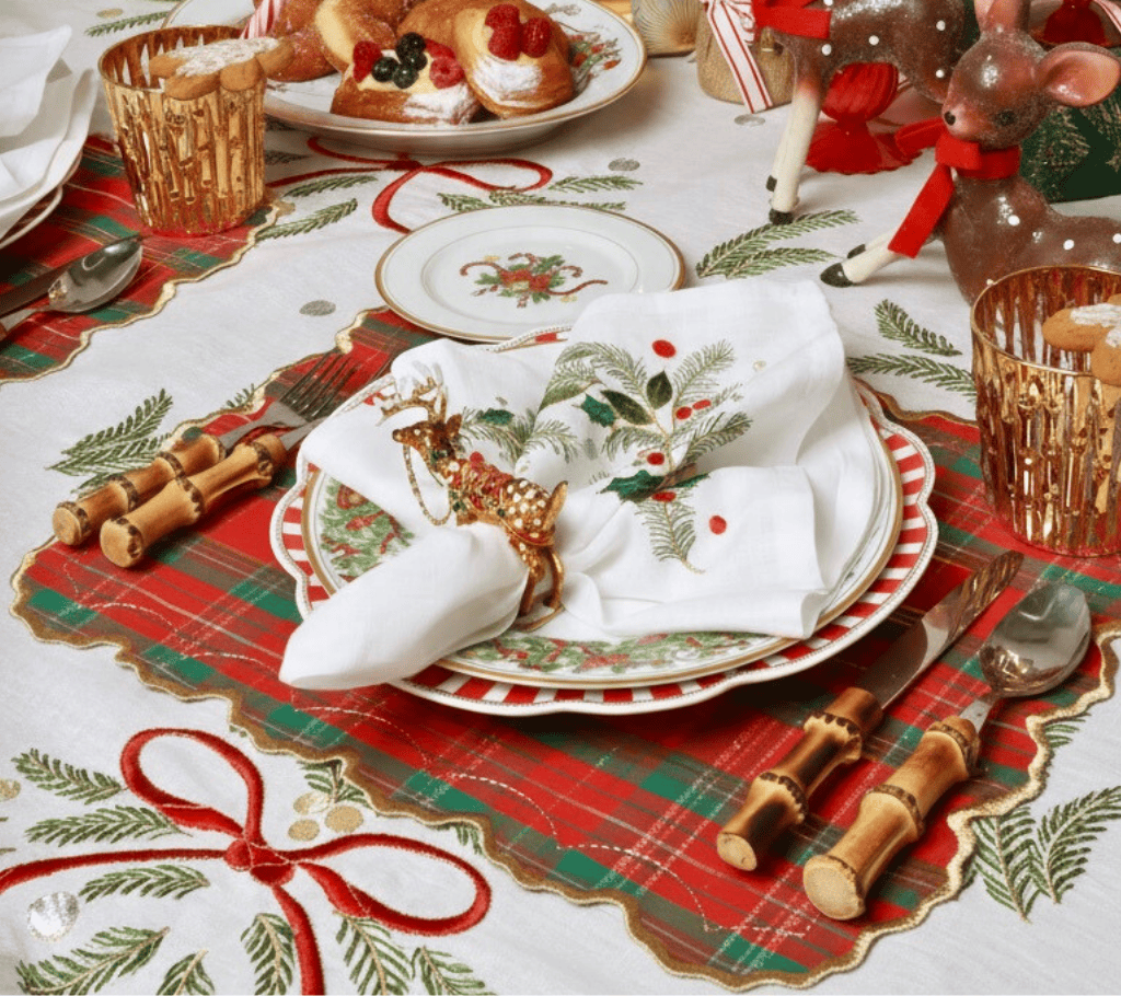 A Merry and Bright Tablecloth by Kim Seybert featuring red ribbons and greenery, set with a plaid placemat, an embroidered holiday napkin, a reindeer napkin ring, and bamboo flatware.
