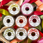Nine spools of Fancy Ribbon in various colors, including a lovely olive ribbon, are arranged in a square pattern and viewed from above. Each spool features a label with a bow illustration and the brand name “Hester & Cook,” making them perfect for gift wrapping.