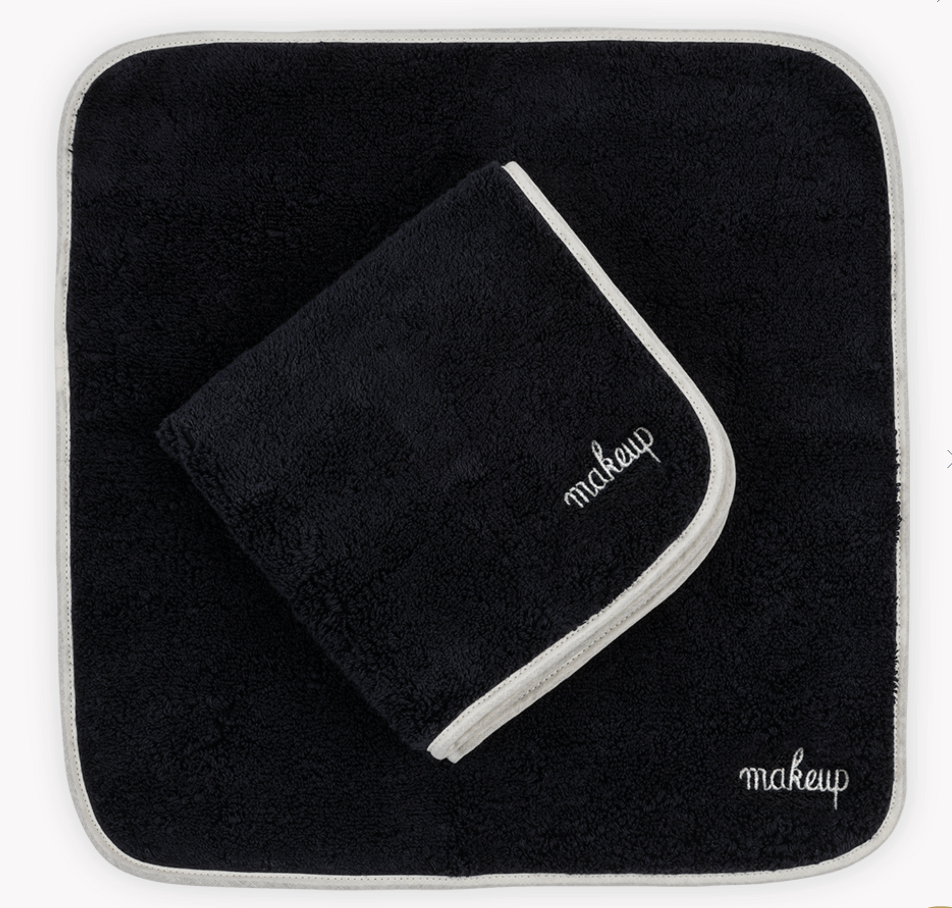 The Matouk Giza Makeup Towel Set features two black, fade-resistant towels with white "makeup" embroidery—one flat, one folded—adding a chic touch to your vanity.