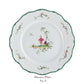 White dinner plate from the Raynaud Longjiang Dinnerware Collection with a scalloped green edge, floral accents, and two figures in traditional attire near trees. Below: "Dinner Plate No. 6". Inspired by Vieux Marseille pastoral scenes.