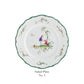 The Raynaud Longjiang Dinnerware Collection salad plate features a white base with a scalloped green rim, floral accents, and two figures by a tree, reminiscent of classic pastoral scenes. Text below reads “Salad Plate No. 5.”.