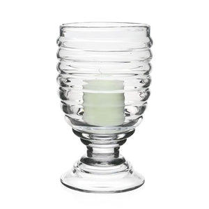 The William Yeoward Crystal Ripples Footed Hurricane is a hand-blown ribbed glass candle holder with a white candle inside, displaying a subtle ripple effect against a plain white background.