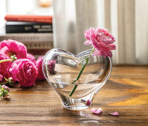 The Simon Pearce Romance Vase, Small, a handmade glass heart-shaped piece, rests on a wooden table holding a pink flower. Additional flowers and stacked books in the background highlight Simon Pearce's signature lead-free glassblowing techniques.