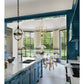 In a modern kitchen featuring blue cabinets and marble countertops, a round dining table is placed by expansive windows overlooking the garden. Gold and glass light fixtures hang from the ceiling, evoking a classic-meets-modern style inspired by bespoke interiors in the spirit of Stewart Manger, as beautifully celebrated in Romancing the Home by Rizzoli.