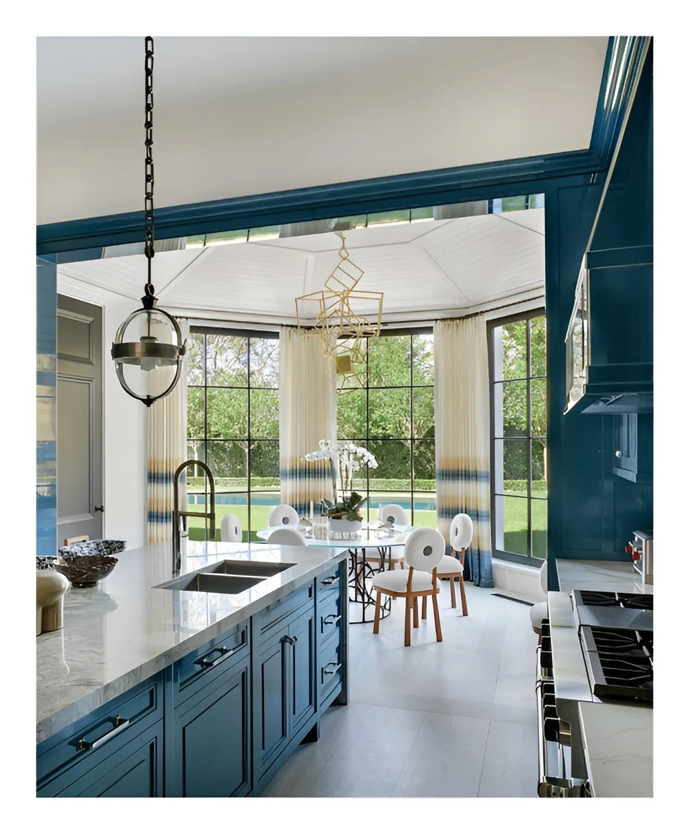 In a modern kitchen featuring blue cabinets and marble countertops, a round dining table is placed by expansive windows overlooking the garden. Gold and glass light fixtures hang from the ceiling, evoking a classic-meets-modern style inspired by bespoke interiors in the spirit of Stewart Manger, as beautifully celebrated in Romancing the Home by Rizzoli.