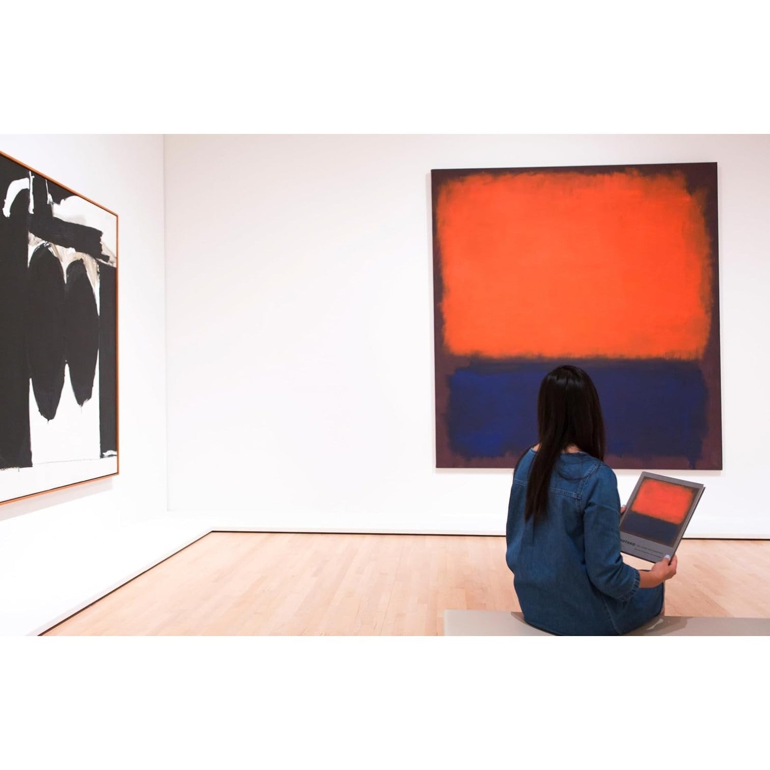 A person sits on a bench in an art gallery with a laptop, facing “Rothko” by Chronicle Books—a striking abstract painting dominated by red and dark hues evocative of classic color field works. Another black and white Abstract Expressionism artwork is displayed on the left wall.