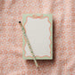 The Rifle Paper Co. Ribbon Notepad with a mint border and a decorative pencil are placed on a pink and white floral patterned fabric background.