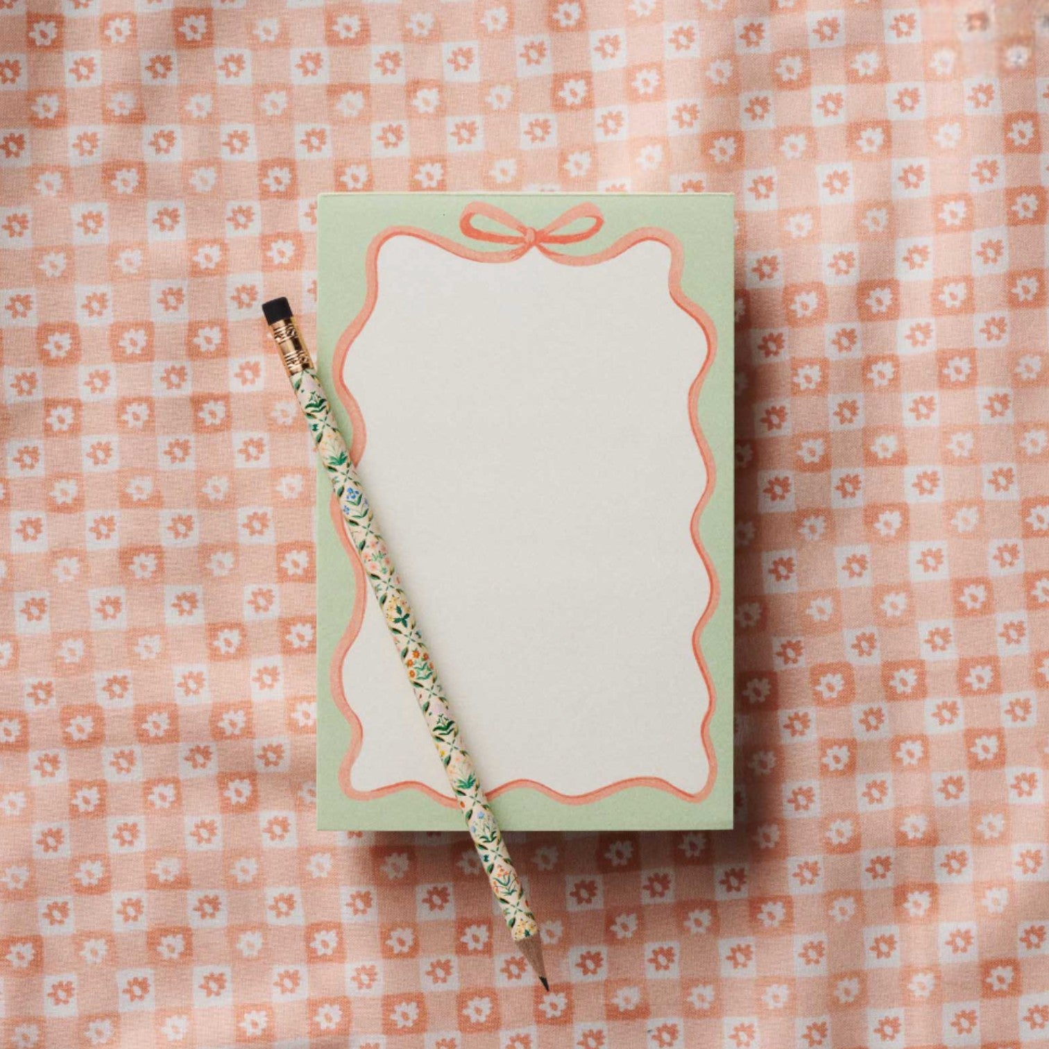 The Rifle Paper Co. Ribbon Notepad with a mint border and a decorative pencil are placed on a pink and white floral patterned fabric background.