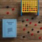 A vertical strategy game board of Connect Four stands upright with red and black checkers scattered around on a wooden floor. A light blue "Connect Four Vintage Bookshelf Edition" box by WS Game Company is placed to the side, adding to its nostalgic charm.