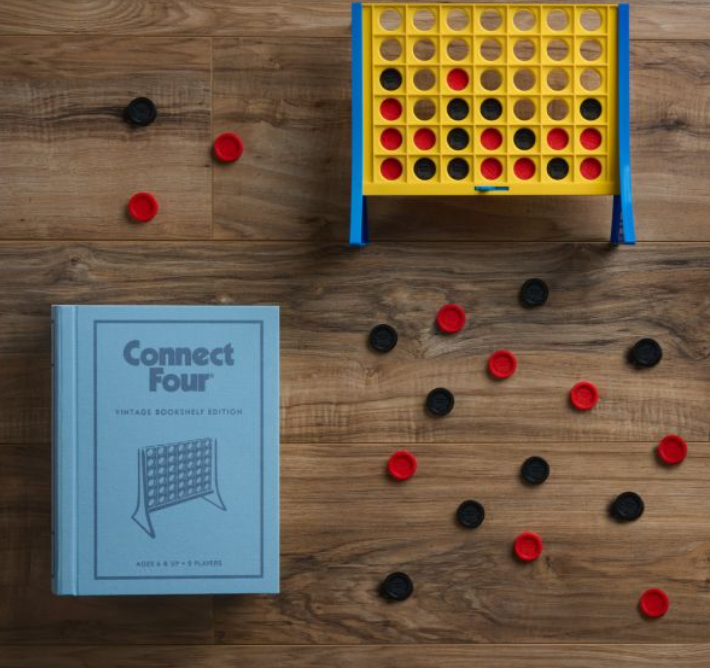 A vertical strategy game board of Connect Four stands upright with red and black checkers scattered around on a wooden floor. A light blue "Connect Four Vintage Bookshelf Edition" box by WS Game Company is placed to the side, adding to its nostalgic charm.
