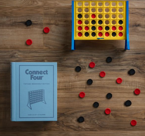 A vertical strategy game board of Connect Four stands upright with red and black checkers scattered around on a wooden floor. A light blue "Connect Four Vintage Bookshelf Edition" box by WS Game Company is placed to the side, adding to its nostalgic charm.