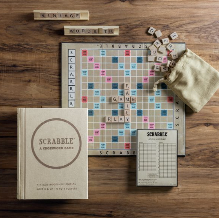 A Scrabble Vintage Bookshelf Edition game setup on a wooden table with wood letter tiles, a scorecard, a fabric-wrapped book tile bag, and the WS Game Company game box, featuring the vintage 1948 game board for players aged 8 and up.