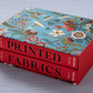 Two red hardcover books, "The Book of Printed Fabrics" by Taschen, explore the history of fabric printing and come in a blue floral slipcase inspired by the Musée de l’Impression sur Étoffes, stacked on a light surface.