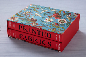Two red hardcover books, "The Book of Printed Fabrics" by Taschen, explore the history of fabric printing and come in a blue floral slipcase inspired by the Musée de l’Impression sur Étoffes, stacked on a light surface.