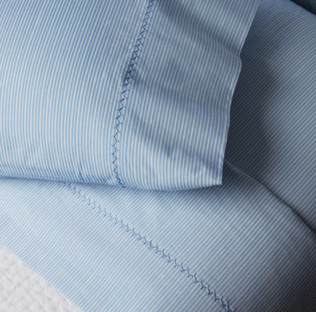 Close-up of the Nandi Light Indigo Sheet Set by John Robshaw, featuring blue and white pinstripes, two pillows, and a blanket with hand-embroidered edges, crafted from organic cotton in India.