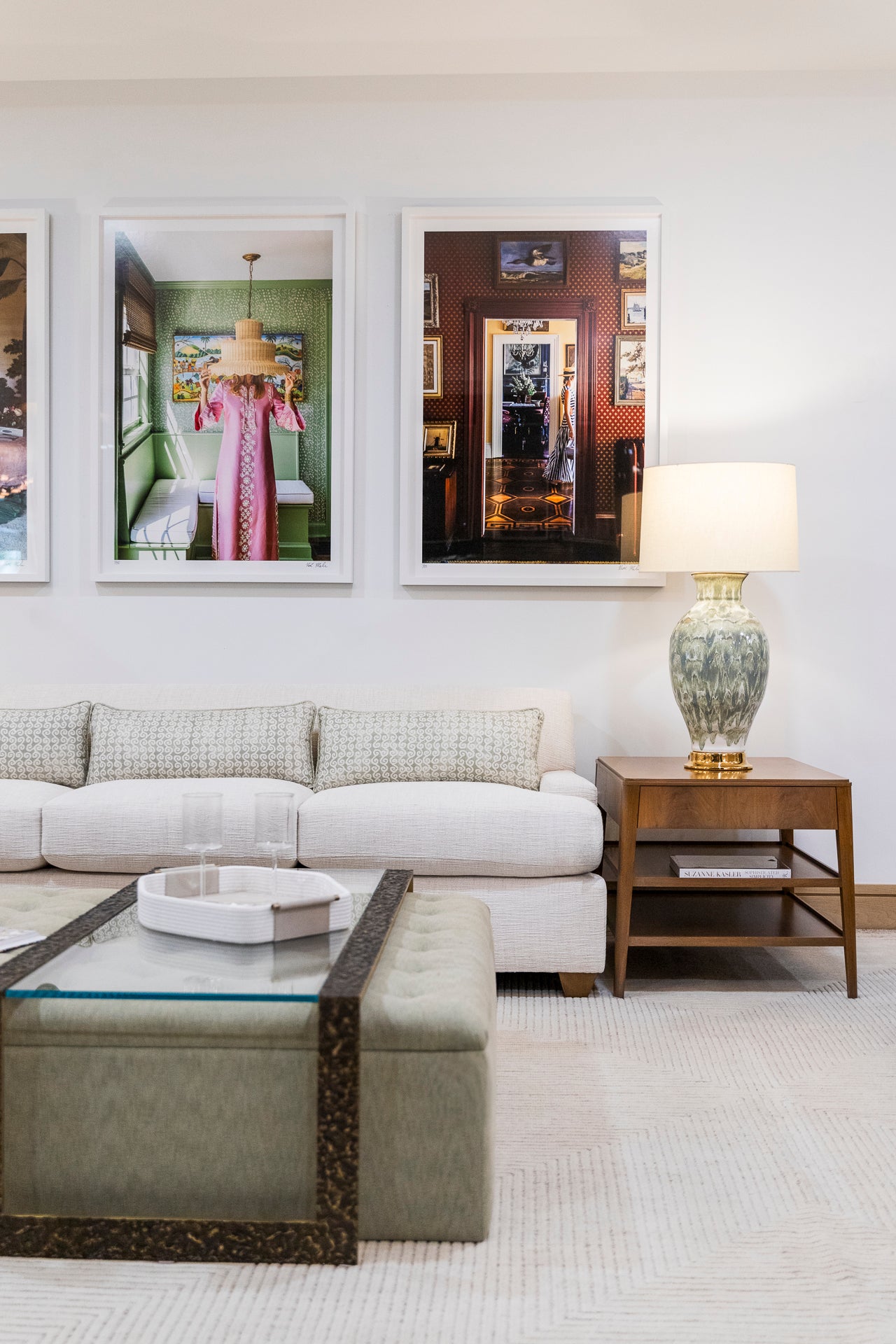 A modern living room with the Baker Yves Sofa, a luxury piece known for its quality, paired with a glass coffee table, green ottoman, lamp on a wooden side table, and framed artwork on the wall.