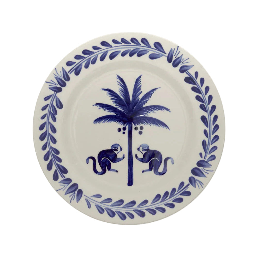The Casa Deep Navy Dinnerware Collection by Johanna Ortiz features a white plate with a blue leafy border, two blue monkeys, and a coconut palm, all handcrafted from Carmen de Viboral clay and expertly hand-painted.