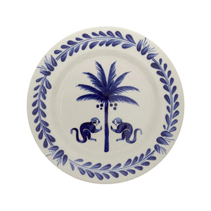 The Casa Deep Navy Dinnerware Collection by Johanna Ortiz features a white plate with a blue leafy border, two blue monkeys, and a coconut palm, all handcrafted from Carmen de Viboral clay and expertly hand-painted.