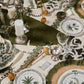 A dining table set with the Casa Dark Jungle Dinnerware Collection by Johanna Ortiz, featuring green and white patterned plates, glassware, utensils, silver candlesticks, and a botanical-themed tablecloth that highlights artisanal heritage.