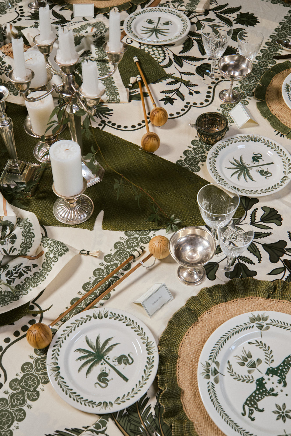 A dining table set with the Casa Dark Jungle Dinnerware Collection by Johanna Ortiz, featuring green and white patterned plates, glassware, utensils, silver candlesticks, and a botanical-themed tablecloth that highlights artisanal heritage.