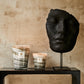 A large black sculpted head on a stand beside two Baobab Collection Sand Atacama candles on a narrow table against a textured beige wall.