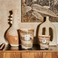 Two rustic ceramic vases and a Baobab Collection Sand Siloli scented candle on a wooden shelf against a wall with a botanical print.