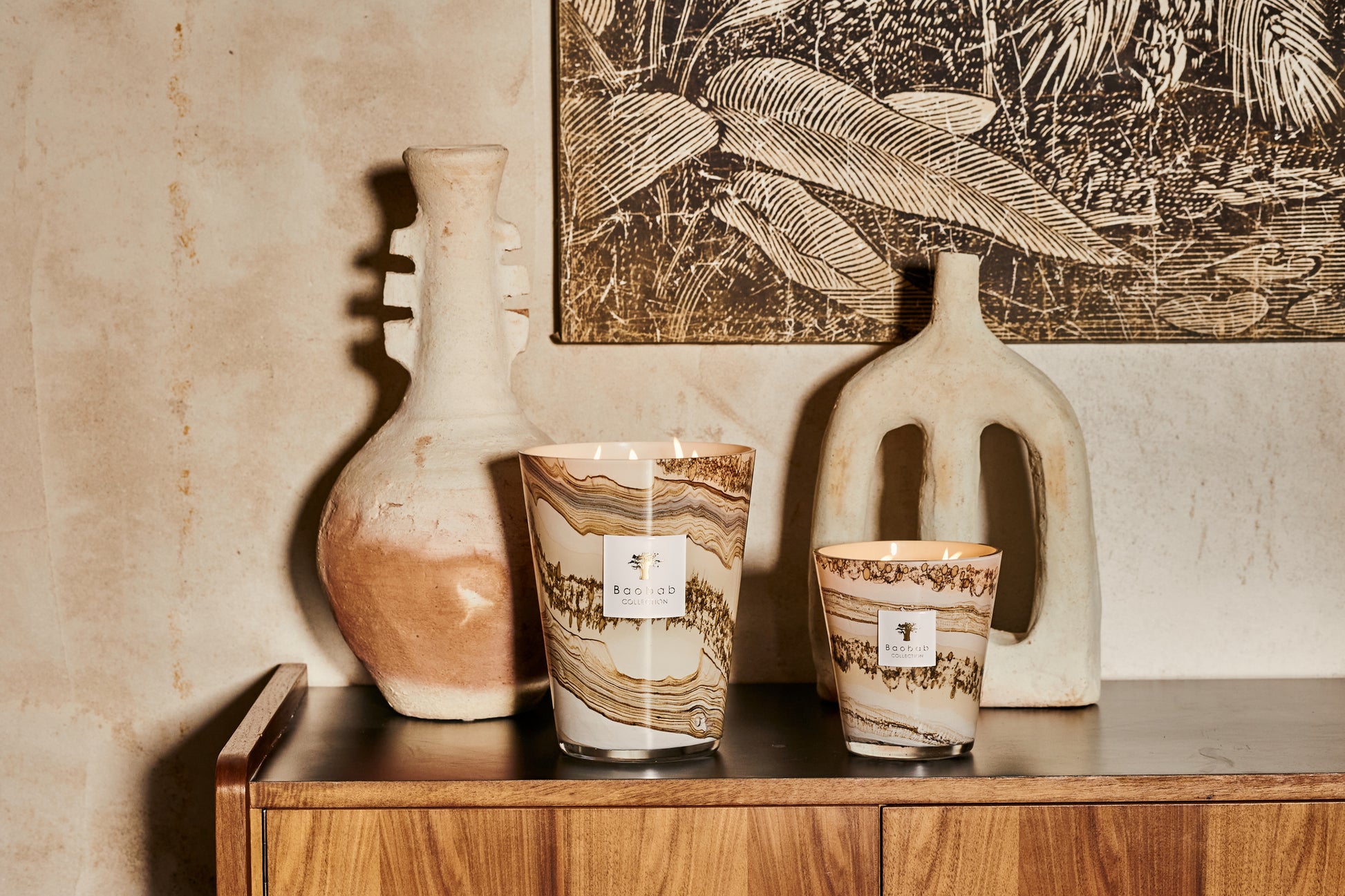 Two rustic ceramic vases and a Baobab Collection Sand Siloli scented candle on a wooden shelf against a wall with a botanical print.