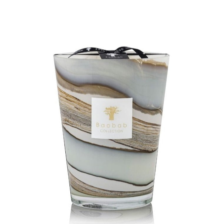 A large decorative candle in a glass container with marbled layers of grey, white, and beige wax. Infused with the exotic scents of myrrh and patchouli, it features a black ribbon with text and a label that reads "Baobab Collection Sand Sonora Candle" attached to the top.