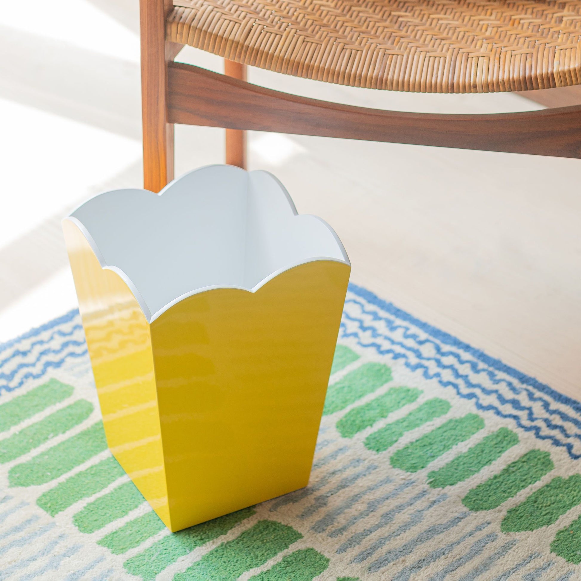 An Addison Ross Scalloped Lacquer Waste Bin in yellow is next to a wooden chair with a woven seat, resting on a colorful rug with green and blue patterns.