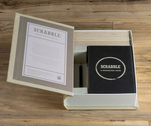 The Scrabble Vintage Bookshelf Edition by WS Game Company is open, revealing its contents including wooden letter tiles and a 1948 vintage-style game board housed inside a fabric-wrapped book. The set is placed on a wooden surface.