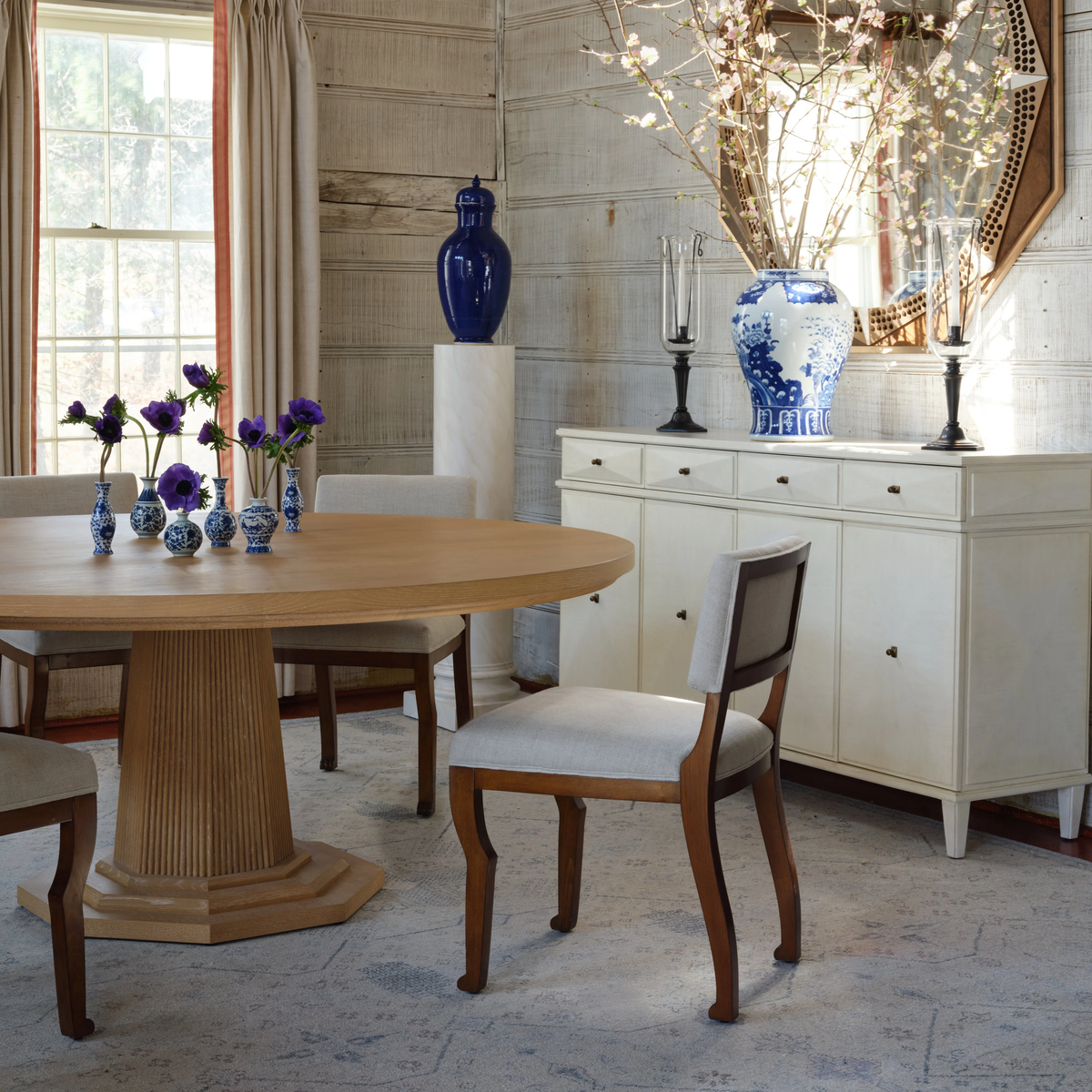 Dining room with the Haven Dining Table by Bunny Williams, a round cerused oak table featuring a classic pedestal base, surrounded by four chairs. Complemented by a white sideboard, blue and white vases, fresh flowers, and a large window letting in natural light.