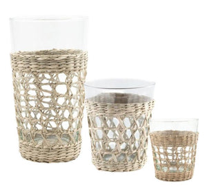 The Seagrass Cage Glassware Collection by Be Home features three clear glass cups in varying sizes, each encased in a light woven seagrass holder—stylish drinkware ideal for serving cocktails.