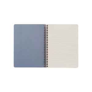 A Seaside Spiral Notebook by Rifle Paper Co., featuring a blue, Seaside-illustrated left cover and a lined right page of heavy-weight textured paper, is shown from above against a white background.