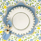 A plate with a blue and white scalloped charger sits on a floral tablecloth, paired with a blue napkin and the Duggan Society Palm Napkin Ring set to the left.