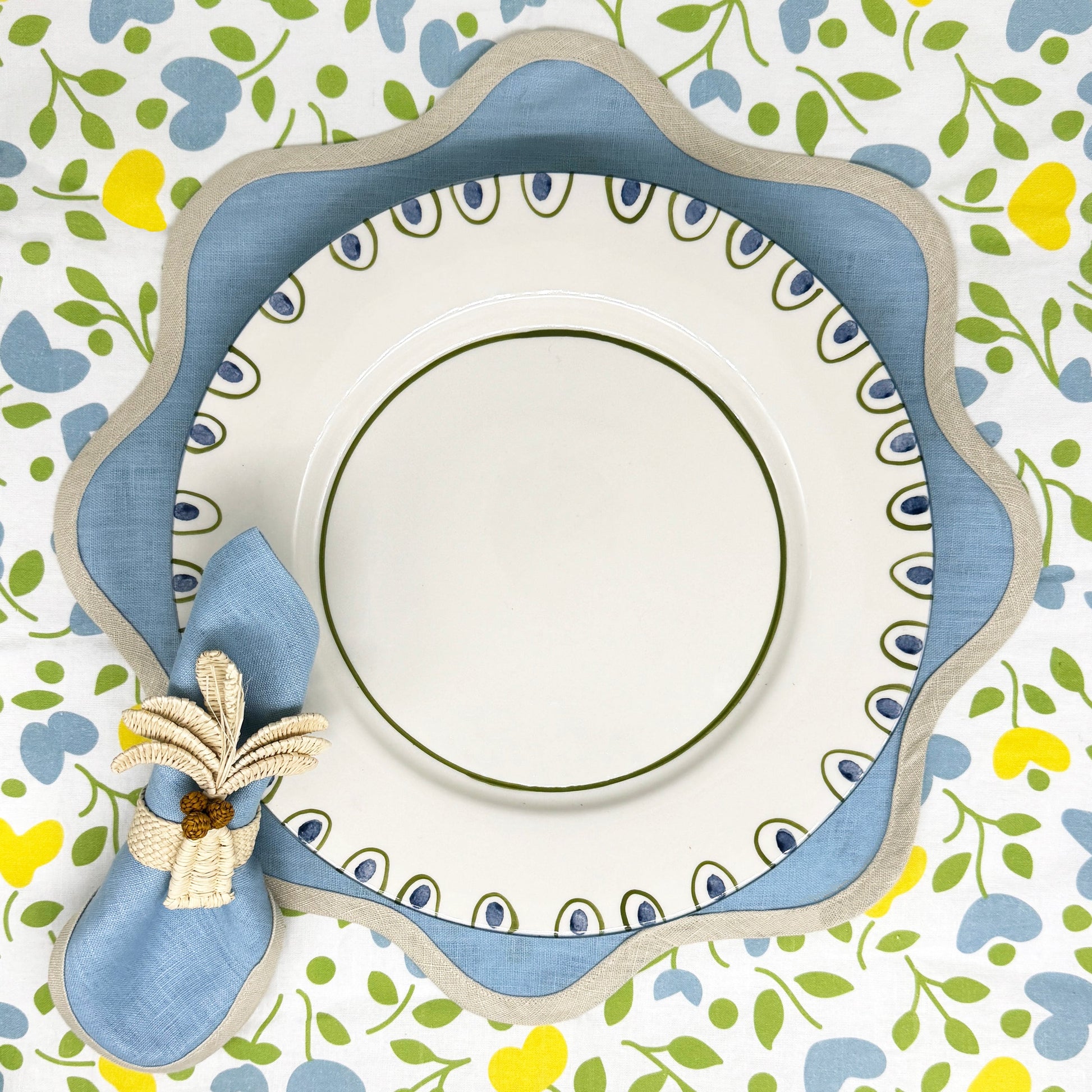 A plate with a blue and white scalloped charger sits on a floral tablecloth, paired with a blue napkin and the Duggan Society Palm Napkin Ring set to the left.