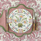 A plate from the Duggan Society Sorrento Dinnerware Collection sits on a pink scalloped placemat with green trim, paired with wicker-handled cutlery, atop a pink and white floral tablecloth for an elegant Italian vibe.