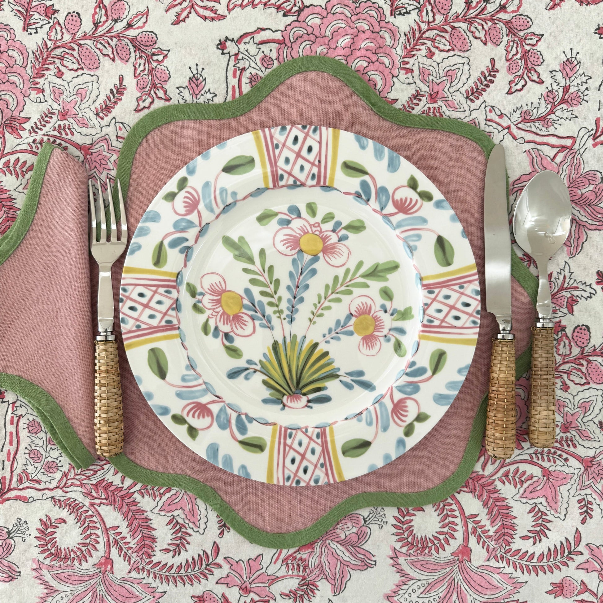 A plate from the Duggan Society Sorrento Dinnerware Collection sits on a pink scalloped placemat with green trim, paired with wicker-handled cutlery, atop a pink and white floral tablecloth for an elegant Italian vibe.