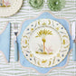 Table set with Duggan Society’s Marina Palms Dinnerware Collection: a decorative botanical plate, blue-edged placemat, matching cutlery, and green glasses on a patterned tablecloth—an elegant coastal touch.