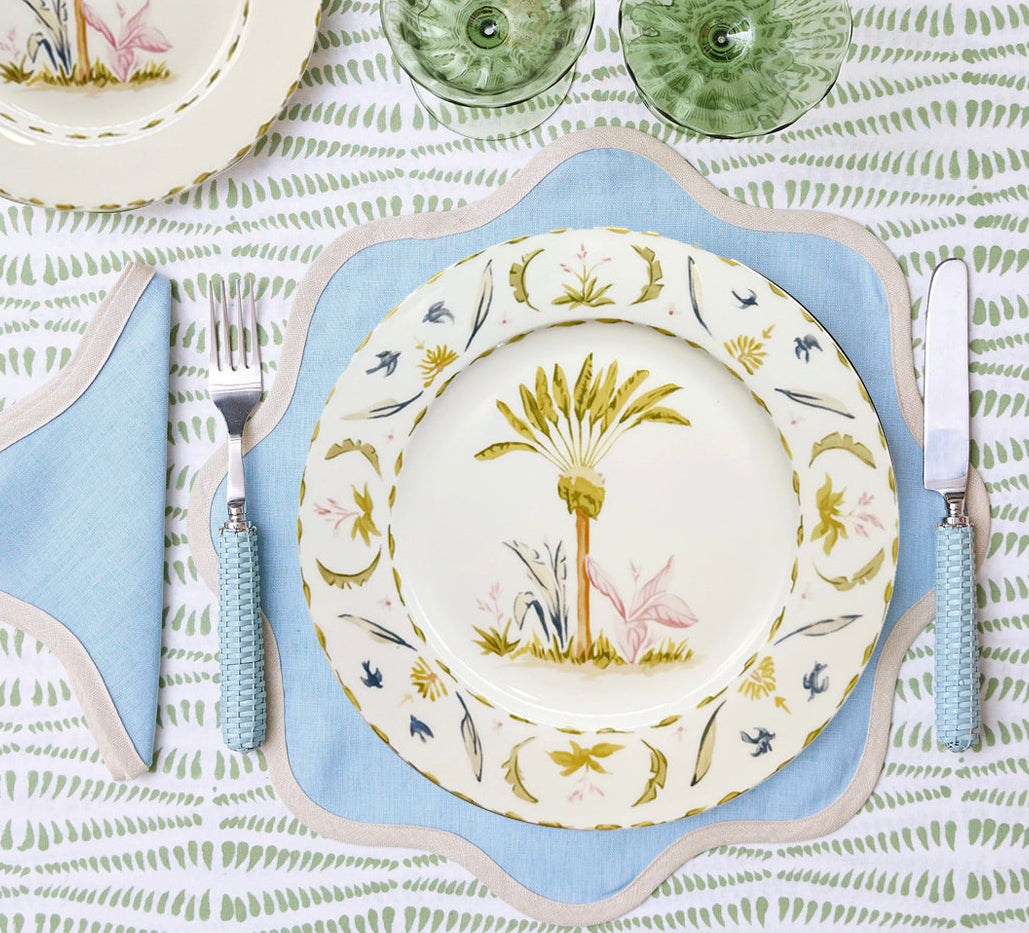 Table set with Duggan Society’s Marina Palms Dinnerware Collection: a decorative botanical plate, blue-edged placemat, matching cutlery, and green glasses on a patterned tablecloth—an elegant coastal touch.
