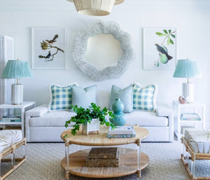 A bright living room with a chalk-white finish, a white sofa adorned with light blue and white checkered pillows, two green lamps, a PALECEK Miramar Mirror featuring a round core rattan design, and bird-themed wall art.